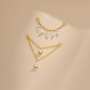 Peut inclure: Deux bijoux dorés. L'un est une chaîne incurvée avec des pierres précieuses claires en forme de goutte d'eau. L'autre est une chaîne triangulaire avec un bijou carré et un bijou en forme de goutte. Le fond est de couleur beige uni.