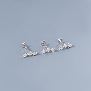 Op de afbeelding: Drie zilverkleurige labret-oorbellen met een platte achterkant en een staaf die zich uitstrekt tot drie heldere, sprankelende zirkonia-stenen. De oorbellen worden gepresenteerd tegen een lichtblauwe achtergrond.