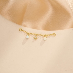 May include: A gold-toned chain connector featuring two round white opal-like beads and a central starburst charm. The delicate metal chain has a circular ring at each end, designed to link two separate jewelry pieces together with a celestial accent.