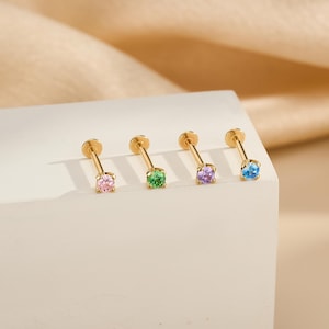 May include: Four gold-colored stud earrings with colorful gemstones. The gemstones are pink, green, purple, and blue. The earrings are displayed on a white surface with a neutral background.