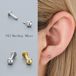 Könnte beinhalten: Silberne und goldfarbene Ohrstecker mit Kugeldesign. Der Text "925 Sterling Sliver" ist sichtbar. Ein Ohrring ist in einem Ohrpiercing abgebildet.