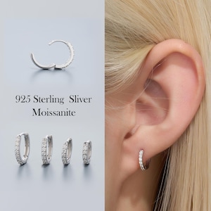 Puede incluir: Pendientes de aro plateados con pequeñas piedras brillantes. La imagen muestra cuatro tamaños diferentes de pendientes, junto con un solo pendiente abierto y un pendiente usado en una oreja. El texto dice "925 Sterling Sliver Moissanite."