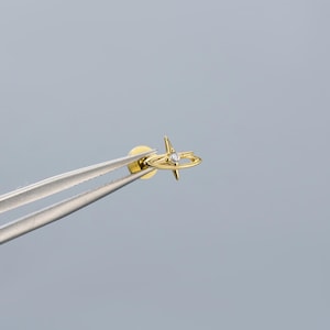 May include: A gold-colored stud earring with a starburst design and a small, clear gemstone at the center. The earring is held by silver tweezers against a light gray background. The earring's design includes a circular band around the starburst.