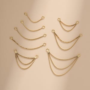 May include: A collection of gold-toned box chains arranged in various configurations, including single, double, and triple draped strands. Each chain segment features small circular jump rings at both ends for attachment, showcasing a metallic finish.