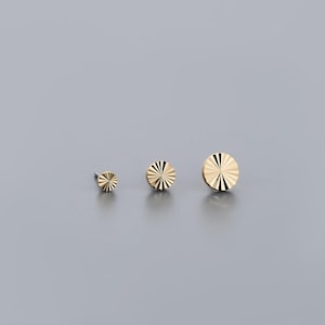 K&ouml;nnte beinhalten: Drei goldfarbene, kreisf&ouml;rmige Ohrstecker in verschiedenen Gr&ouml;&szlig;en. Jeder Ohrring hat ein Sonnenstrahl-Design mit radialen Linien. Die Ohrringe werden auf grauem Hintergrund pr&auml;sentiert und zeigen ihre polierten, reflektierenden Oberfl&auml;chen.