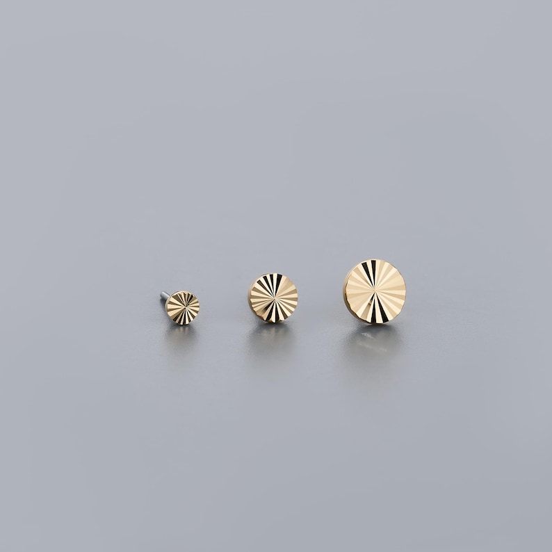 K&ouml;nnte beinhalten: Drei goldfarbene, kreisf&ouml;rmige Ohrstecker in verschiedenen Gr&ouml;&szlig;en. Jeder Ohrring hat ein Sonnenstrahl-Design mit radialen Linien. Die Ohrringe werden auf grauem Hintergrund pr&auml;sentiert und zeigen ihre polierten, reflektierenden Oberfl&auml;chen.