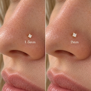 Può includere: Un piercing da naso in oro con una pietra preziosa rotonda e trasparente montata su quattro griffe. L'immagine mostra due opzioni di misura: un piercing da 1,5 mm a sinistra e uno da 2 mm a destra, con le dimensioni indicate sotto ciascuno.