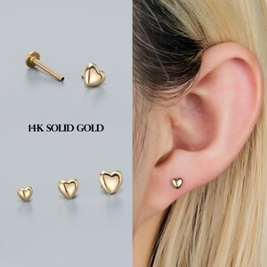 May include: A selection of 14K solid gold heart-shaped earrings in different sizes. One earring is shown in the ear. The earrings are presented against a light grey backdrop. The text "14K SOLID GOLD" is printed in black.