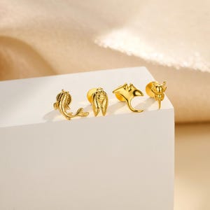 May include: Four gold-colored stud earrings with aquatic and insect designs. The earrings include a koi fish, a cicada, a manta ray, and a beetle. The earrings are displayed on a white surface.