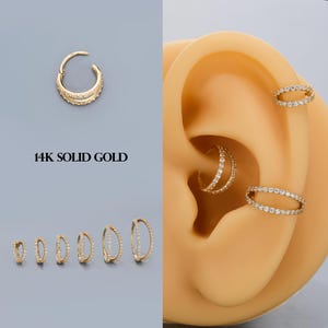 Könnte beinhalten: Goldfarbene Ohrringe mit klaren Edelsteinen. Das Bild zeigt verschiedene Stile, darunter Creolen und offene Kreisdesigns, einige mit doppelten Steinreihen. Der Text "14K SOLID GOLD" ist ebenfalls sichtbar.