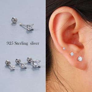 May include: Three pairs of silver stud earrings with clear cubic zirconia stones. The earrings are shown on a model's ear. The earrings are available in different sizes.