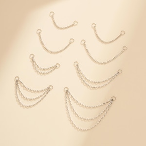 Könnte beinhalten: Silberfarbene Kettenaccessoires. Das Bild zeigt mehrere Ketten unterschiedlicher Längen, einige mit mehreren Strängen. Jede Kette hat an jedem Ende einen kleinen Ring. Die Ketten sind auf einer hellbeigen Oberfläche angeordnet.