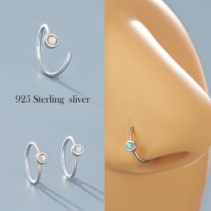 May include: A close-up of a silver nose ring with a small white opal gemstone. The ring is shown on a model's nose and also on a white background.