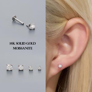 Puede incluir: Una colección de pendientes de moissanita de oro macizo de 14 quilates en varios tamaños. La imagen muestra un solo poste de pendiente, un pendiente de botón y una oreja que lleva un pendiente de botón. También se ve el texto "14K SOLID GOLD MOISSANITE".