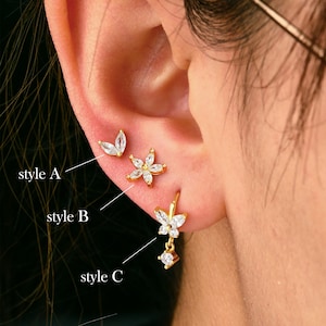 May include: Three styles of earrings with gold settings and clear cubic zirconia stones. Style A is a pair of small, leaf-shaped earrings. Style B is a pair of small, flower-shaped earrings. Style C is a single earring with a dangling flower charm.