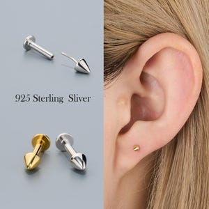 Puede incluir: Pendientes de botón plateados y dorados con diseño de cono. Un pendiente se muestra en la oreja. El texto "925 Sterling Sliver" aparece en la imagen.