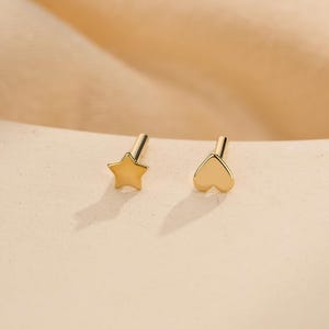 Puede incluir: Un par de pendientes de botón en tono dorado con forma de estrella de cinco puntas y forma de corazón. Cada pendiente tiene un acabado liso y pulido con un poste recto que se extiende desde la parte posterior, diseñado para perforaciones.