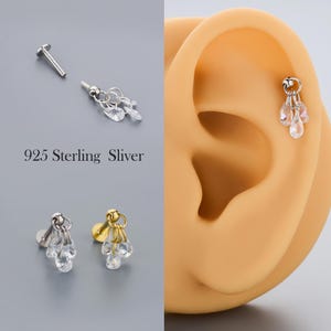 May include: A silver earring with a dangling teardrop design is shown next to an ear. The earring has a small post and a cluster of clear, faceted teardrop-shaped gems. The text "925 Sterling Sliver" is also visible.