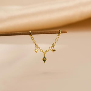 May include: A gold chain earring featuring three star-shaped charms and a green gemstone pendant. The earring hangs from a wooden dowel against a neutral backdrop. The chain is fine, and the charms are intricately designed.