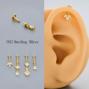 Può includere: Orecchini in argento sterling 925 color oro con pietre di zirconia cubica trasparente. L'immagine mostra vari stili di orecchini, tra cui borchie e modelli pendenti. Il testo "925 Sterling Sliver" è visibile.