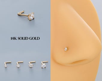 Puede incluir: Una colección de piercings de nariz de oro macizo de 14 quilates con una gema transparente de talla redonda. La imagen muestra los piercings en varios tamaños y un primer plano de uno en una nariz. También se ve el texto "14K SOLID GOLD".