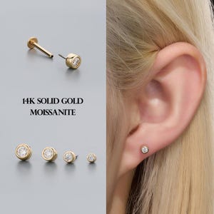 May include: Gold stud earrings with a round, bezel-set moissanite stone. The image shows multiple sizes of the earrings, along with the text "14K SOLID GOLD MOISSANITE". One earring is shown in an ear piercing.