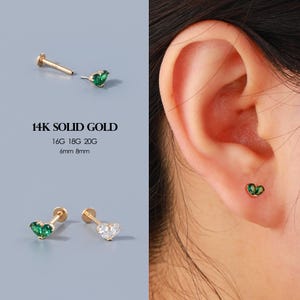 May include: Gold stud earrings with heart-shaped green gemstones. The image shows the earrings in different views, including one worn on an ear. Text on the image reads "14K SOLID GOLD", "16G 18G 20G", "6mm 8mm".