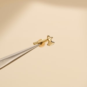 May include: A small, gold-colored axe-shaped earring. The earring is held by tweezers against a neutral background. The axe head is detailed, and the handle has a textured design. The earring is likely made of metal.