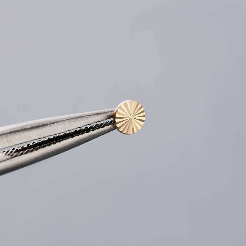 K&ouml;nnte beinhalten: Ein kleiner, goldfarbener, kreisf&ouml;rmiger Ohrstecker mit einem Sonnenstrahl-Design, der von einer Pinzette gehalten wird. Der Ohrring hat eine helle, gl&auml;nzende Goldfarbe. Die Pinzette ist silberfarben und hat einen strukturierten Griff. Der Hintergrund ist einfarbig hellgrau.