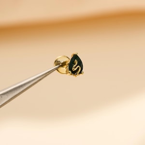Pode incluir: Um brinco de pino com parte traseira plana em ouro, apresentando uma pedra preciosa verde-escura em forma de gota. A pedra é acentuada com um fio dourado fino em forma de serpente que se curva sobre a superfície. Base circular e pino.
