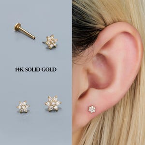 Puede incluir: Un conjunto de pendientes de oro macizo de 14 quilates con forma de flor y gemas transparentes. Se muestra un pendiente en la oreja. El texto "14K SOLID GOLD" se muestra en la imagen.