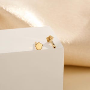 May include: Two small, gold-coloured flower-shaped earrings. One earring is a stud, and the other has a post. The earrings are on a white surface with a gold background. The earrings are likely made of gold or a gold-coloured metal.