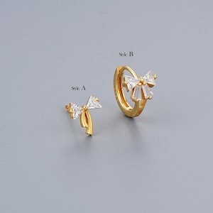 May include: Two gold bow earrings with clear cubic zirconia stones. The earring on the left is a stud and the earring on the right is a hoop. The text "Style A" is above the stud earring and the text "Style B" is above the hoop earring.