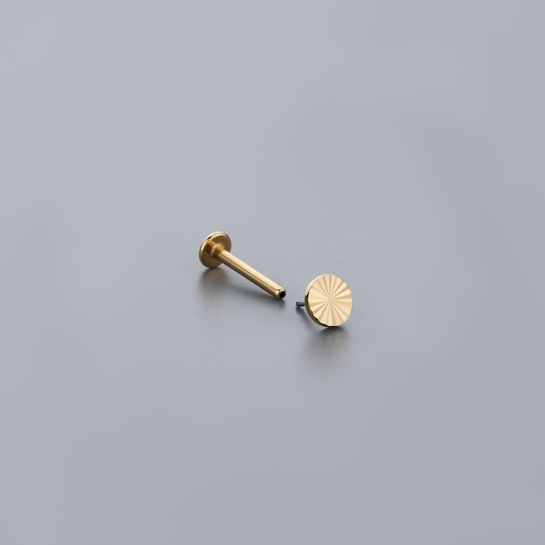 K&ouml;nnte beinhalten: Zwei goldfarbene Ohrringe mit flachem R&uuml;cken. Ein Ohrring ist ein Stecker mit einer flachen, runden Scheibe. Der andere Ohrring ist ein Stecker mit einer runden, strukturierten Scheibe mit Sonnenstrahlendesign. Die Ohrringe sind auf grauem Hintergrund.