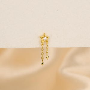 May include: A gold-colored earring featuring a star-shaped top with a clear gemstone in the center. Two delicate chains dangle from the star, each ending in a small, geometric charm. The earring is set against a soft, neutral background.