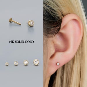 Könnte beinhalten: Goldene Ohrstecker mit einem eingefassten, klaren Edelstein. Das Bild zeigt verschiedene Größen der Ohrringe, zusammen mit dem Text "14K SOLID GOLD". Ein Ohrring ist in einem Ohr-Piercing zu sehen.
