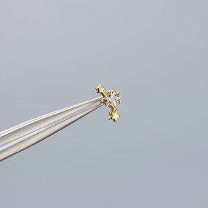 May include: A gold-colored earring with a star design. The earring features a central clear gemstone star, with two smaller gold stars dangling below. The earring is held by silver tweezers against a gray background.