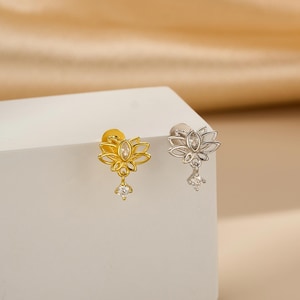 May include: A pair of stud earrings, one gold-toned and one silver-toned, each featuring a lotus flower design. The earrings have a dangling, teardrop-shaped, clear gemstone. The earrings are displayed on a white surface against a soft, neutral background.