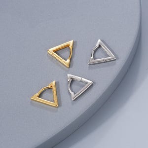 Puede incluir: Cuatro pendientes de aro triangulares, dos plateados y dos dorados. Los pendientes están sobre una superficie gris.