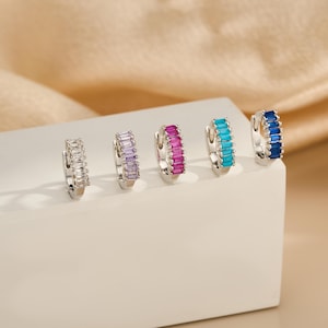 May include: Five silver hoop earrings, each adorned with a row of rectangular gemstones in various colors: clear, purple, magenta, turquoise, and blue. The earrings are displayed on a white surface against a soft, neutral background.
