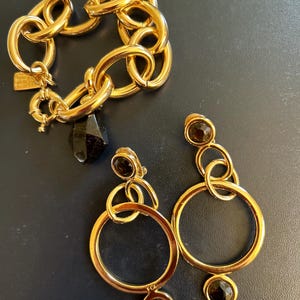 Könnte beinhalten: Ein Set aus goldfarbenem Schmuck mit einem dicken Kettenarmband mit einem schwarzen Steinanhänger und einem Paar Ohrhängern mit schwarzen Steinen und runden Akzenten.