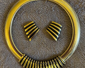 May include: A gold-toned necklace and matching earrings set. The necklace is a thick, ribbed collar with a clasp closure. The earrings are geometric, with black and gold striped designs. The jewelry set is displayed on a textured, gray surface.