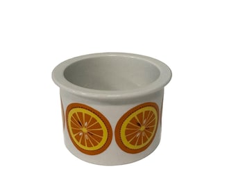 May include: A small, white ceramic container with a wide rim. The container is decorated with repeating orange and yellow citrus fruit slice designs. The interior of the container is a light gray color.