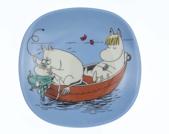 Op de afbeelding: Een blauw keramiek bord met een cartoonillustratie van twee witte Moemins in een rode boot. Eén Moemin bestuurt de boot met een groene motor, terwijl de andere Moemin in de boot ontspant.