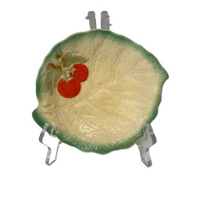 May include: A leaf-shaped ceramic plate with a green rim and a light yellow centre. The plate features a design of two red tomatoes with green leaves. The plate is displayed on a clear plastic stand.