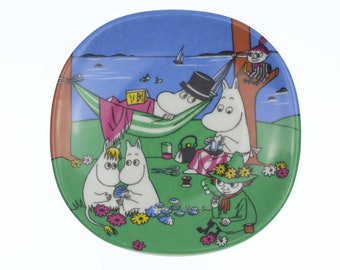 Op de afbeelding: Een wit keramiek bord met een groene rand heeft een kleurrijke illustratie van Moomin-personages die genieten van een picknick. De scène toont een Moomin die ontspant in een hangmat, een andere Moomin die naait en een derde Moomin-personage die zit met een pijp. Het bord heeft een blauwe lucht en water op de achtergrond.