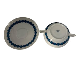 Może przedstawiać: Biały ceramiczny zestaw do zupy i spodek. Miska ma dwa uchwyty, a spodek jest okrągły. Oba elementy zdobione są powtarzającym się wzorem niebieskich owalnych kształtów wokół krawędzi. Miska jest odwrócona do góry nogami.