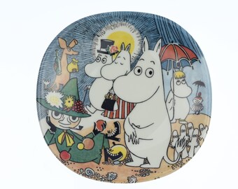 Op de afbeelding: Een witte keramische plaat met een kleurrijke illustratie van Moomin-personages op een strand. Het ontwerp bevat Moomintrolls, Snufkin en andere personages onder een zon en een regenscène. De plaat heeft een blauwe rand.
