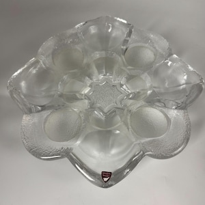 Puede incluir: Fuente de vidrio transparente con diseño floral. La fuente presenta un exterior texturizado y un centro en forma de estrella. El logotipo de Orrefors Sweden es visible en la parte inferior. La fuente tiene múltiples hendiduras circulares.