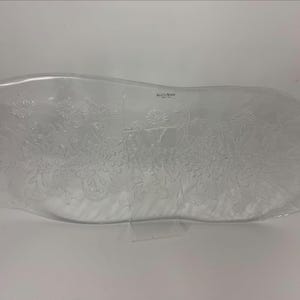 Puede incluir: Fuente ovalada de vidrio transparente con diseño floral. La fuente tiene un borde ligeramente elevado y está decorada con flores y follaje en relieve. El nombre de la marca es visible en la parte superior central. Una pieza decorativa para servir alimentos.